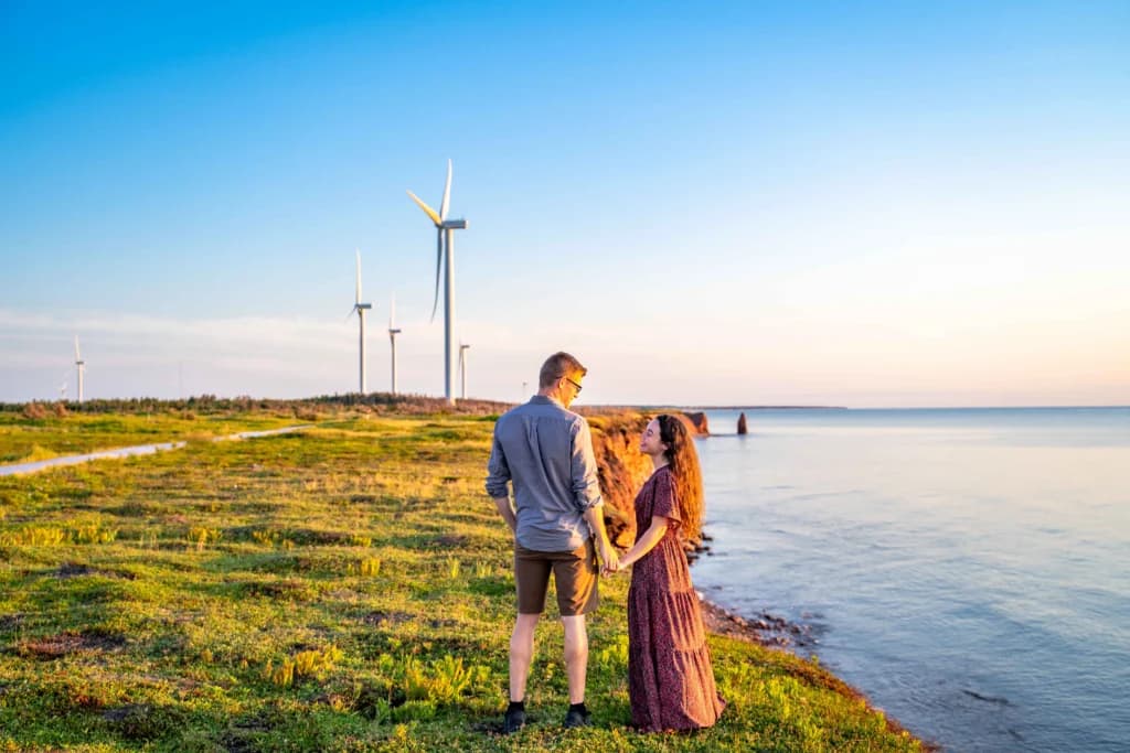 A couple standing on a grassy headland at golden hour, looking out over the water with wind turbines behind them
